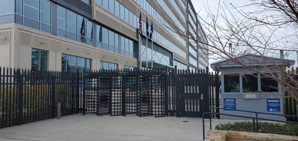 Guardhouse and fence with locked pedestrian gates and large office building in background.